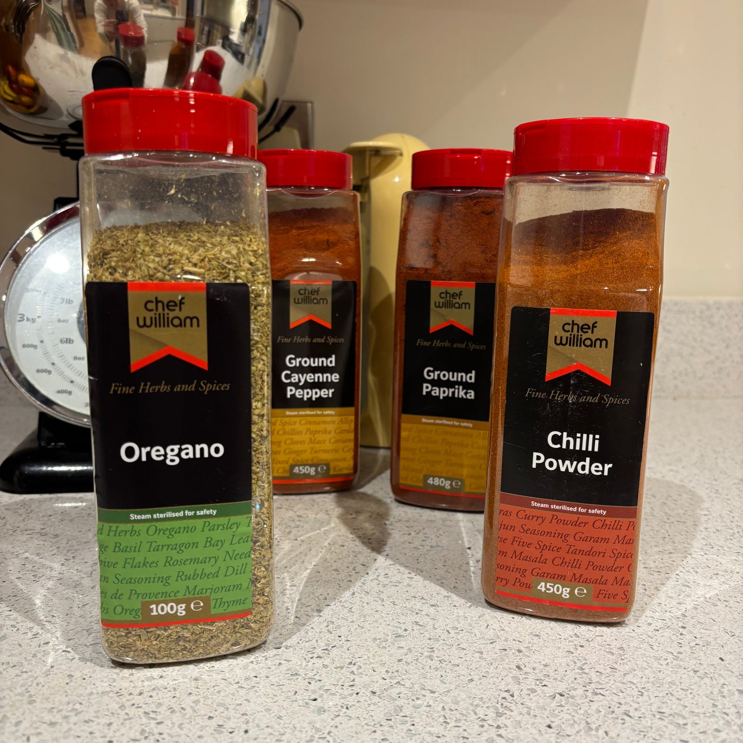 Spice bottles including Chilli Powder, Ground Paprika, Ground Coriander Pepper, and Oregano on a countertop.