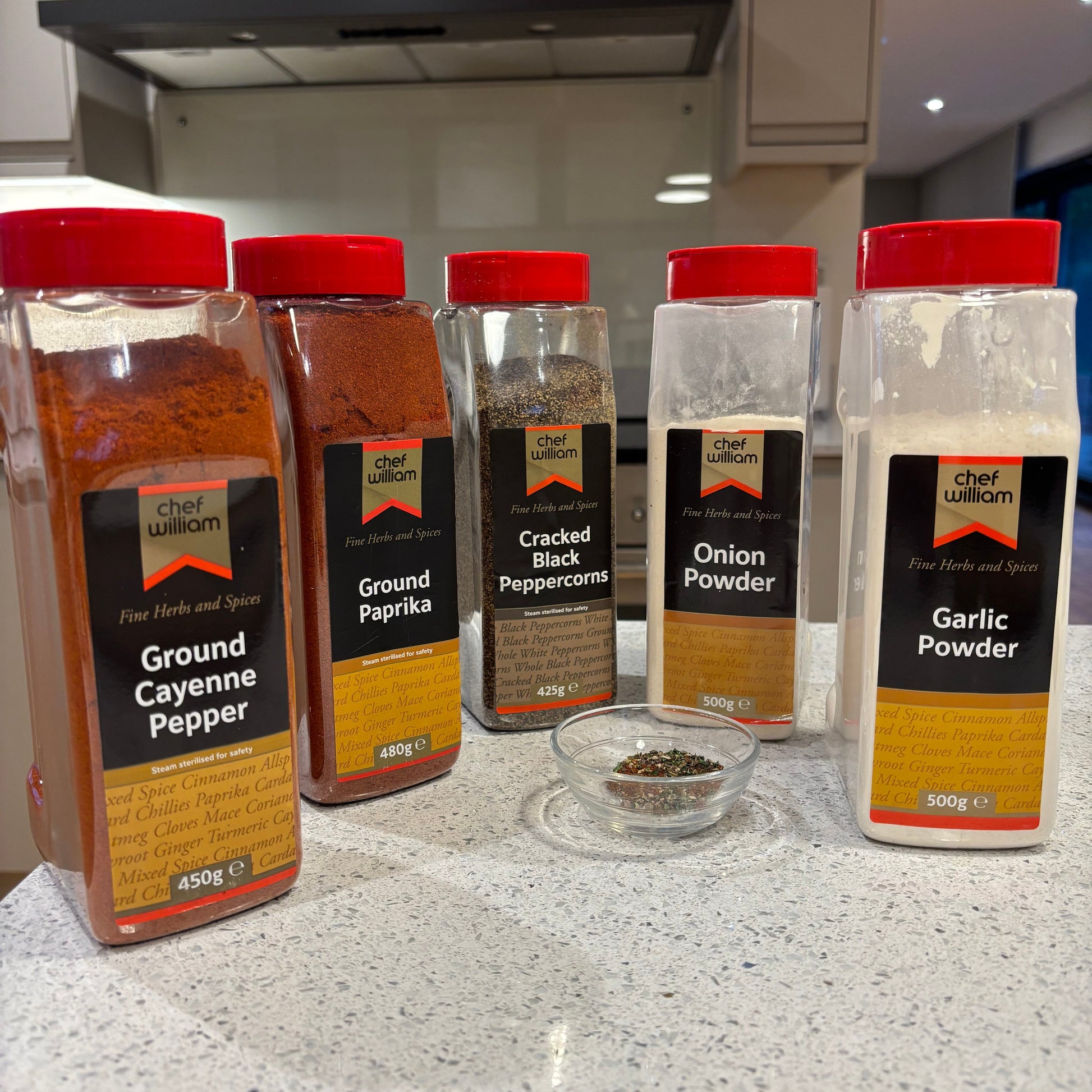 Spice jars including garlic powder, onion powder, cracked black pepper, ground paprika, and cayenne pepper on a countertop with a small bowl of mixed BBQ herbs in front. 